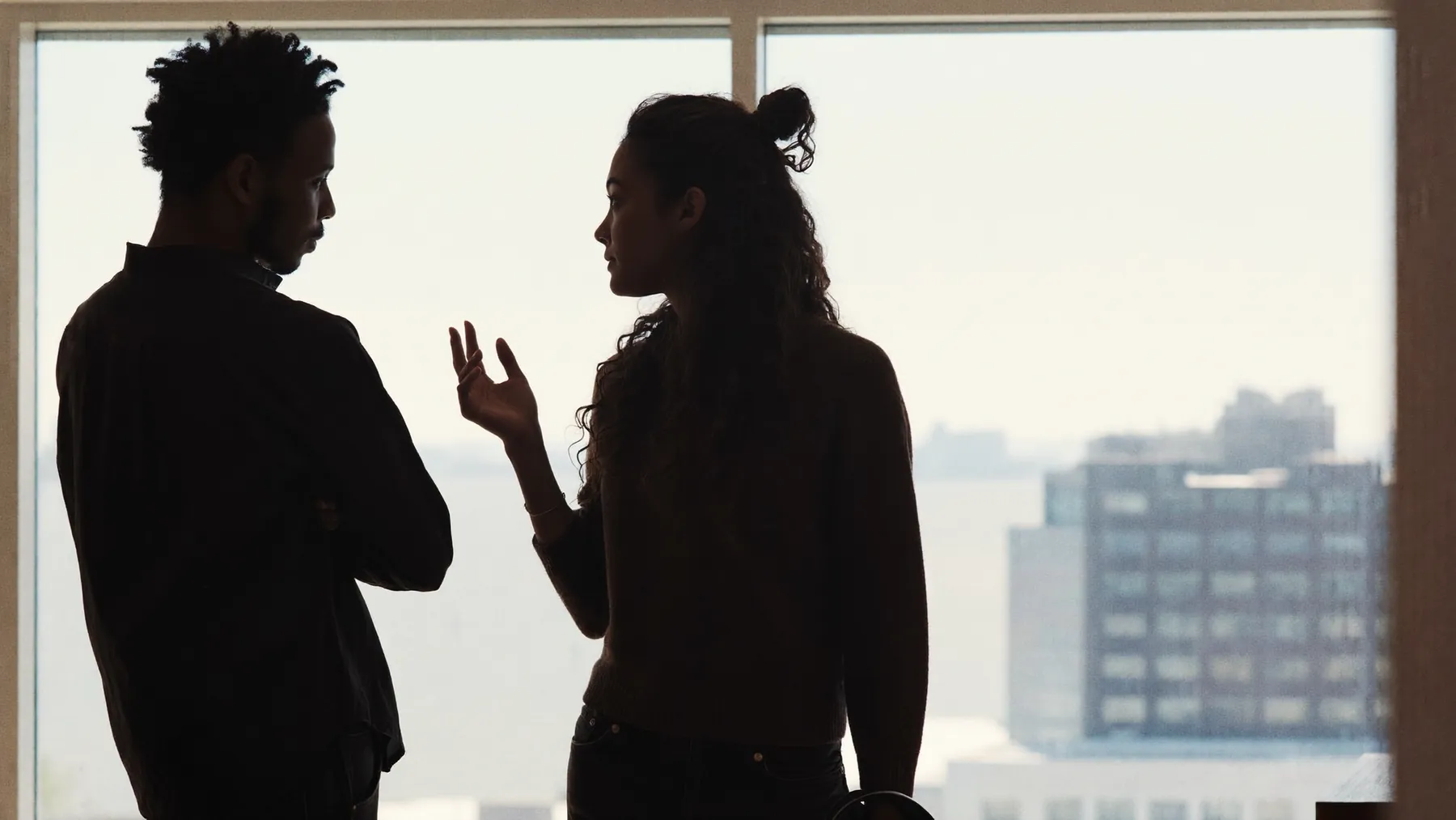 Two people in near-silhouette talking against a bright floor-to-ceiling window